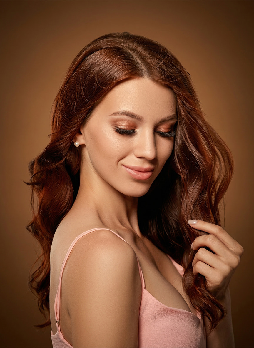 Kobieta z&nbsp;przymkniętymi oczami, dotykająca długich, falowanych, rudych włosów. Zbliżenie prezentujące profesjonalny efekt przedłużania włosów w&nbsp;salonie Rokoko.